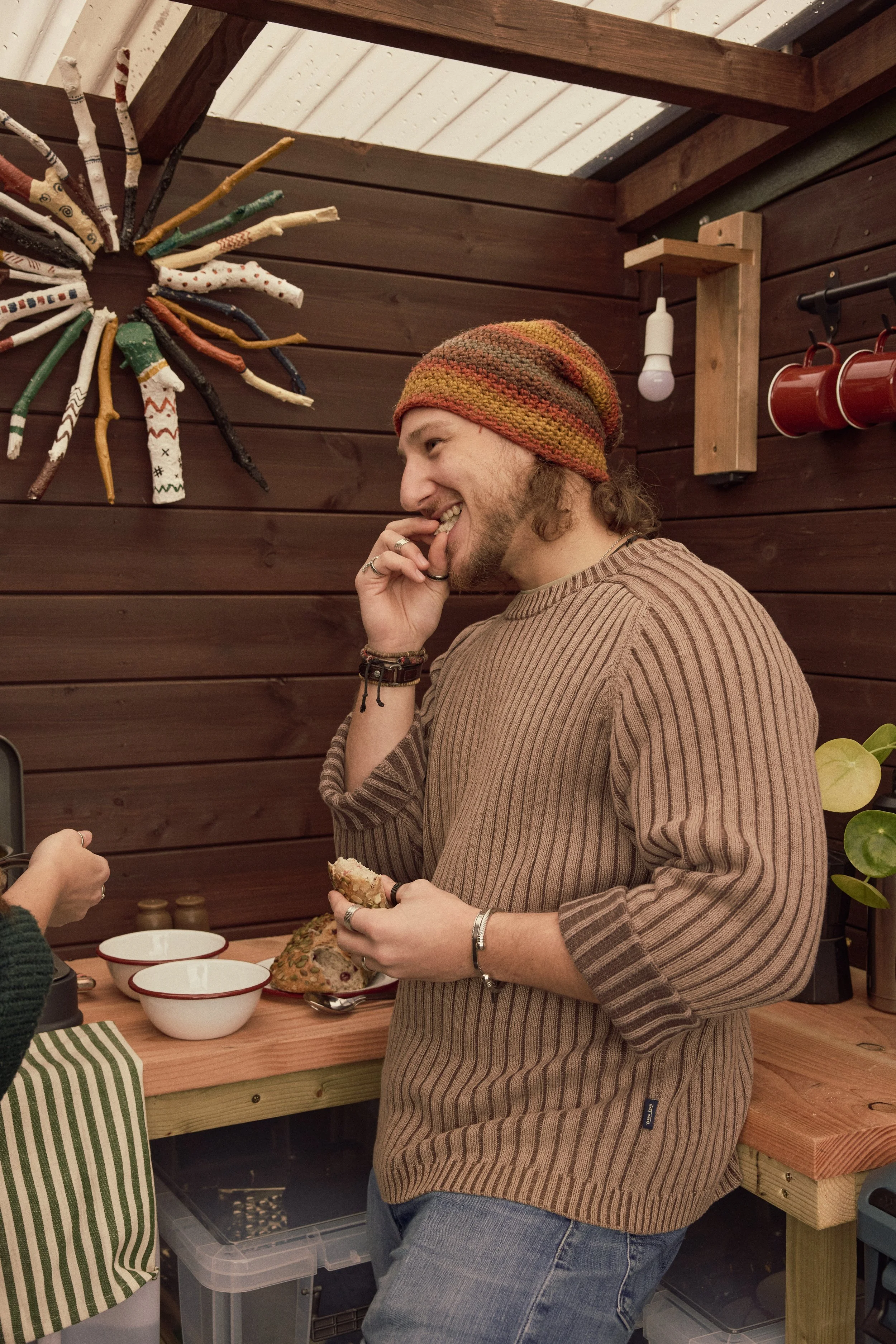 A man with long hair and a beard, wearing a multicolored knit beanie and a striped brown sweater, smiling and touching his lips in a rustic kitchen or workshop. There is a colorful starburst decoration on the wooden wall behind him, with hanging red mugs and an exposed light bulb. The man is holding a slice of bread or cake, and there are bowls and a loaf on the table in front of him.