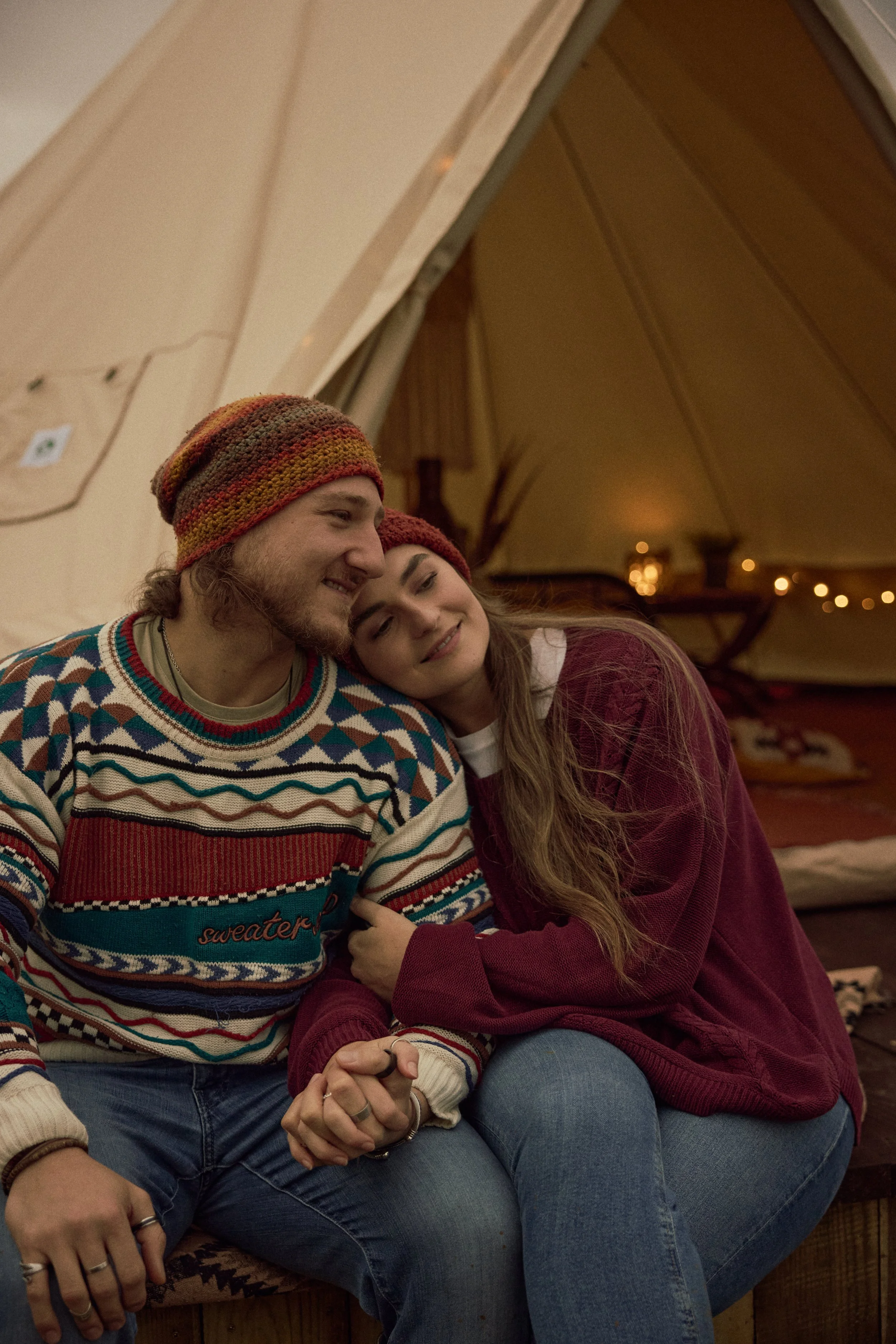 Two young adults, a man and a woman, sitting closely together indoors. The man is wearing a colorful, patterned sweater and a multicolored knit beanie. The woman has long brown hair, is resting her head on the man's shoulder, and is wearing a maroon sweater. They appear happy and comfortable, with the background softly lit and decorated with small string lights.