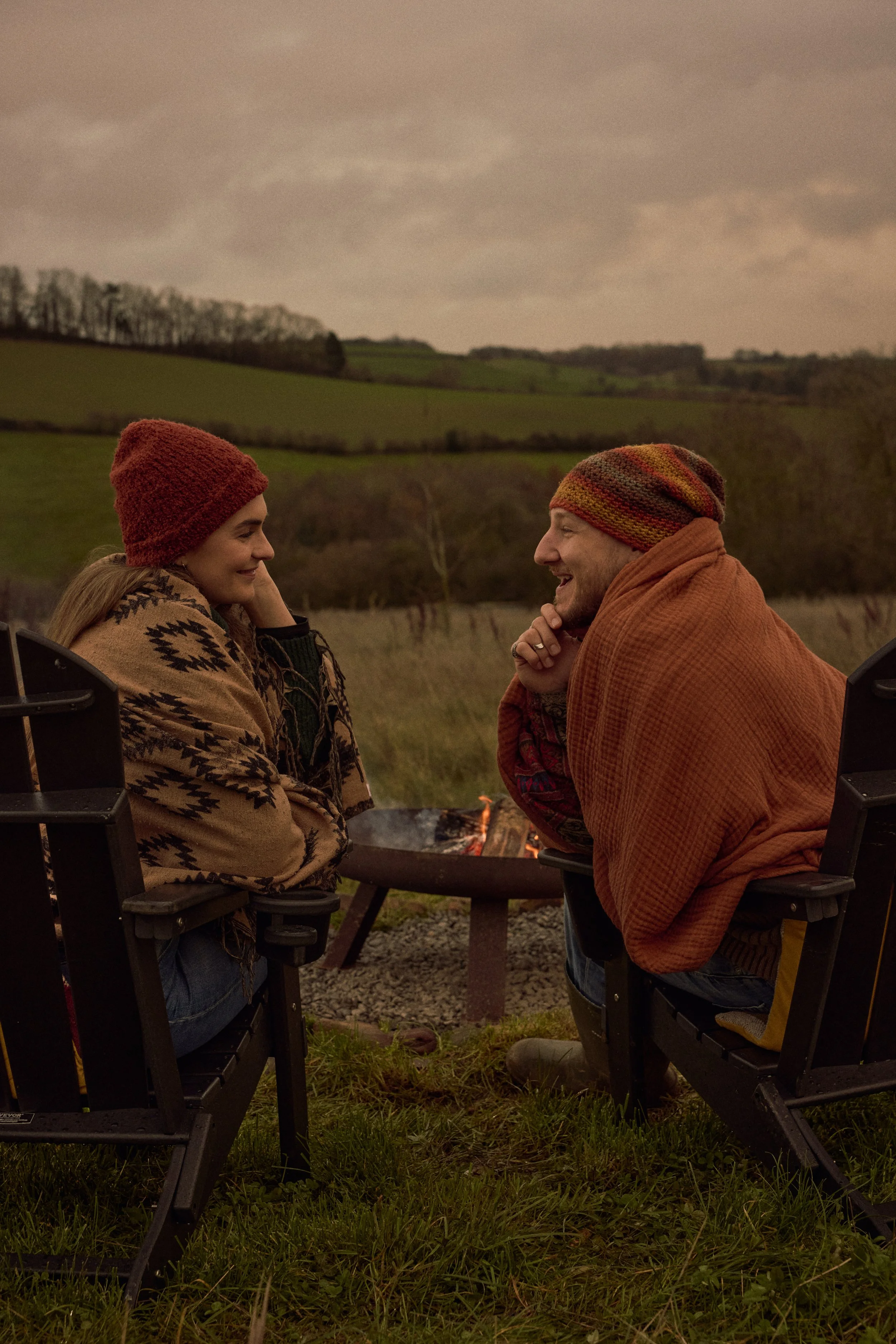 Two people sitting outdoors near a fire, smiling and engaging in conversation with a scenic green landscape and cloudy sky in the background.