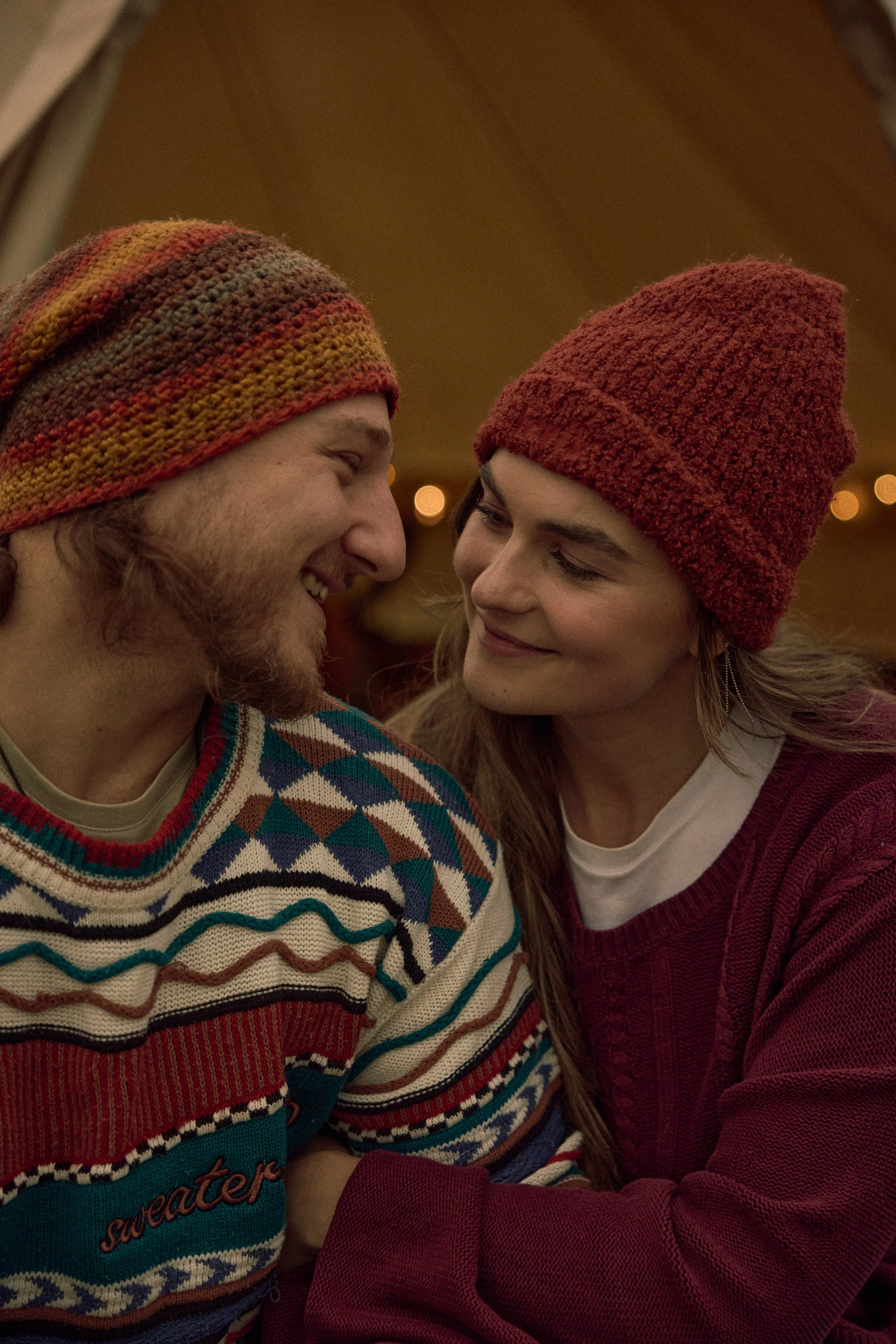 A young man and a young woman are face to face, smiling and looking at each other. They are both wearing warm knit hats, the man's hat has colorful stripes and the woman's is solid red. The man is wearing a patterned sweater, and the woman is wearing a red cardigan over a white shirt. The background is softly blurred with warm, out-of-focus lights.