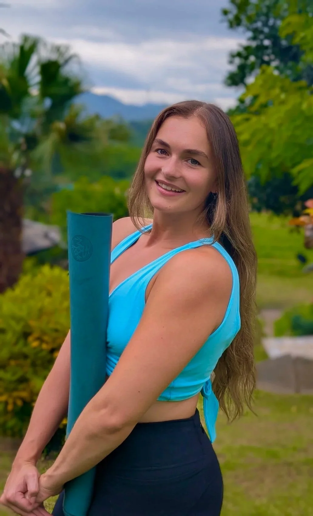 A young woman with long brown hair, wearing a light blue sleeveless top, holding a blue yoga mat outdoors in a lush green garden with trees and mountains in the background.