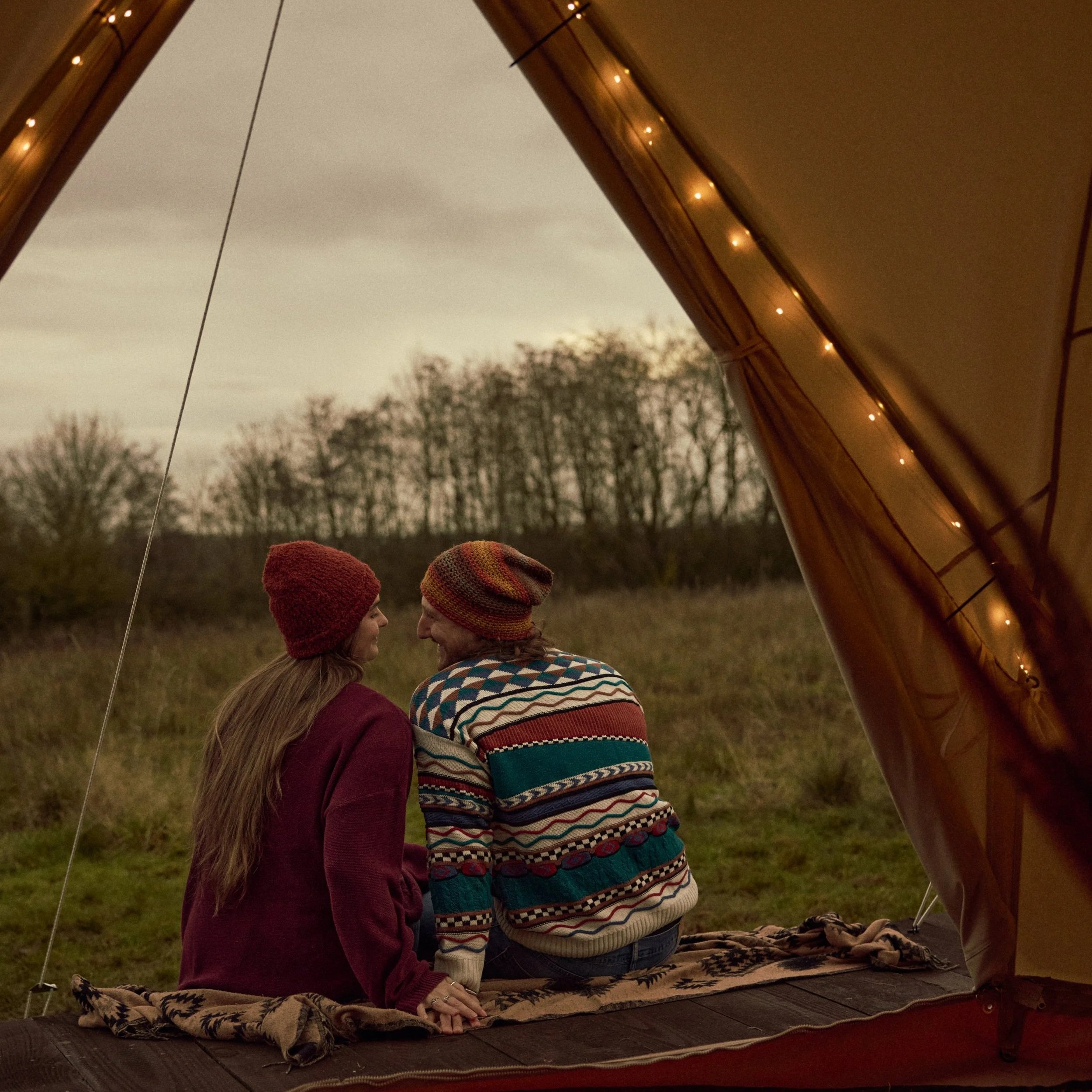 Two people standing inside a tent outdoors at dusk, facing each other, one woman and one man both wearing knit hats, with trees visible in the background.