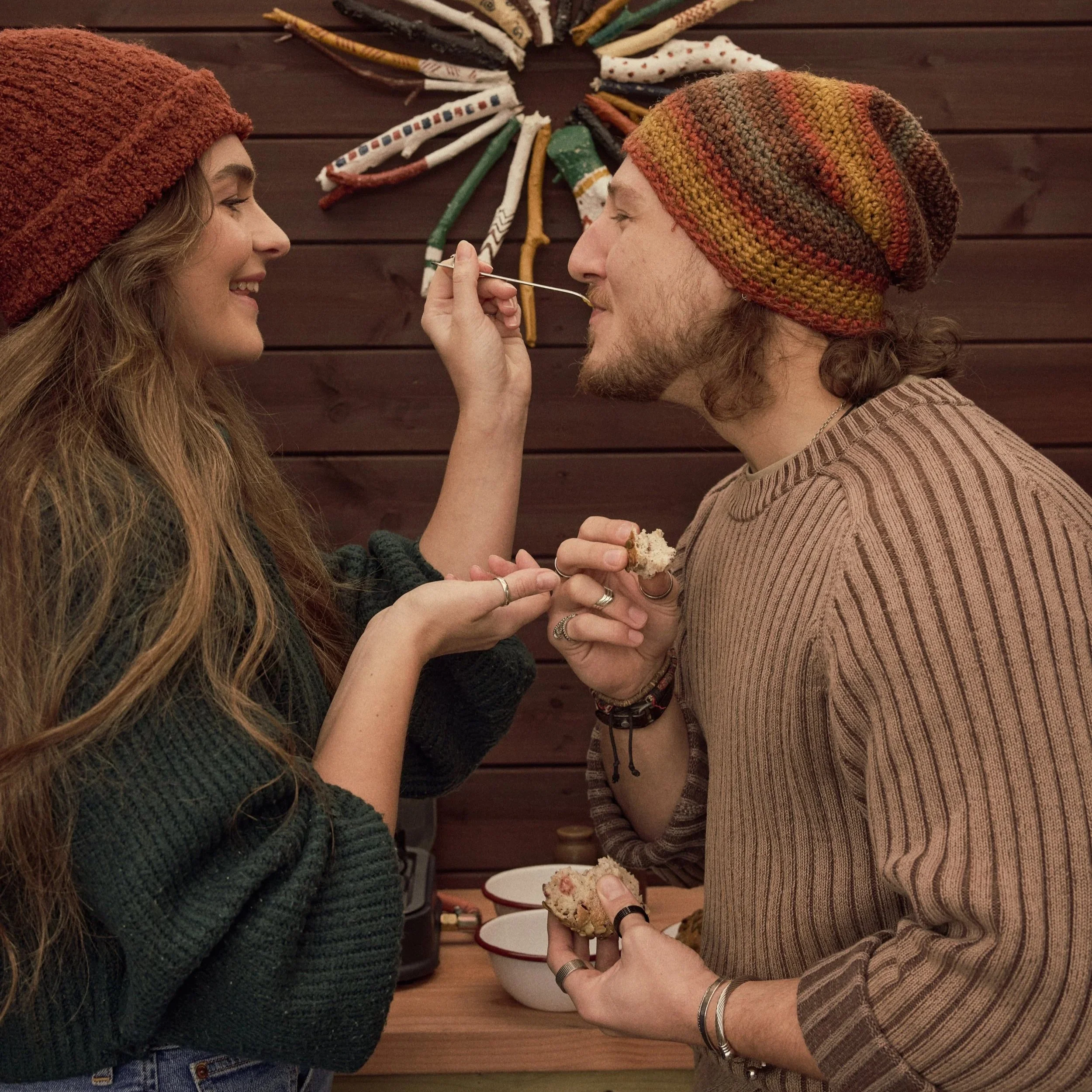 A woman with long wavy hair wearing a dark green knitted sweater and a rust-colored knit hat, smiling and facing a man. The man has long curly hair, facial hair, and is wearing a multicolored knitted hat and a striped beige shirt. holding a piece of cake and a fork, offering a bite.