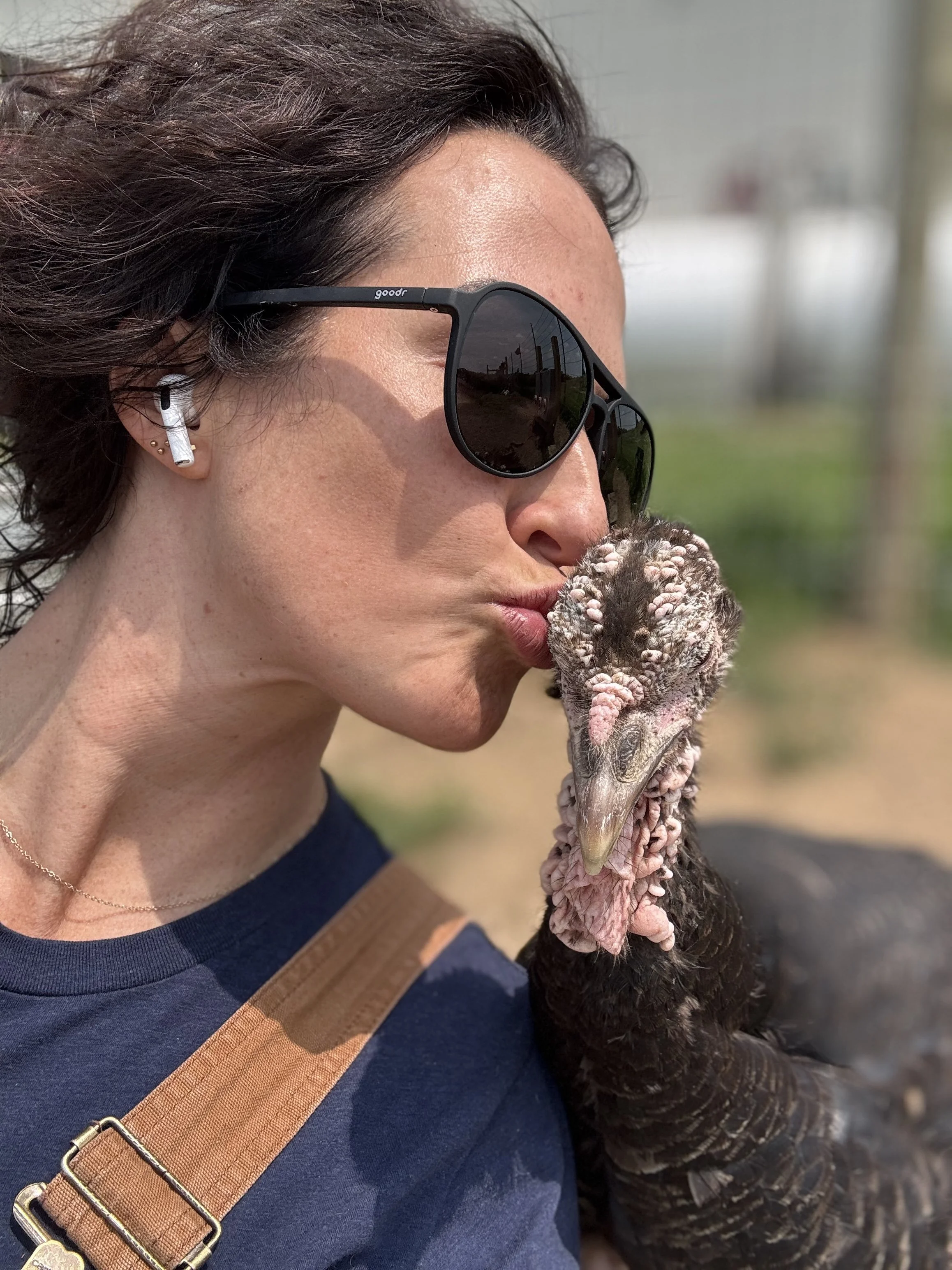 A woman with dark curly hair, wearing black sunglasses, AirPods, and a navy blue shirt, is kissing a turkey.