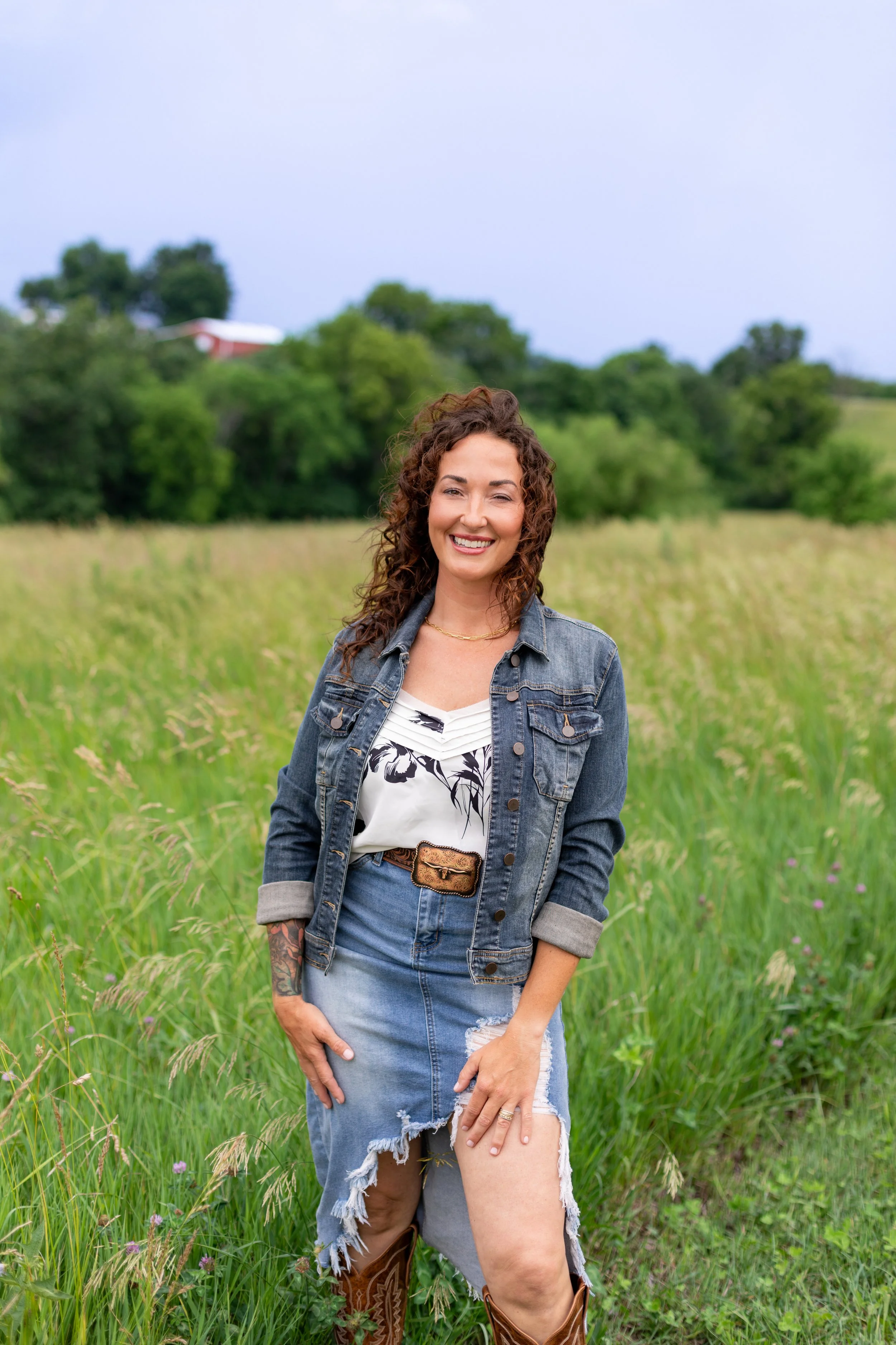A woman smiling and standing in a grassy field with trees and a cloudy sky in the background.