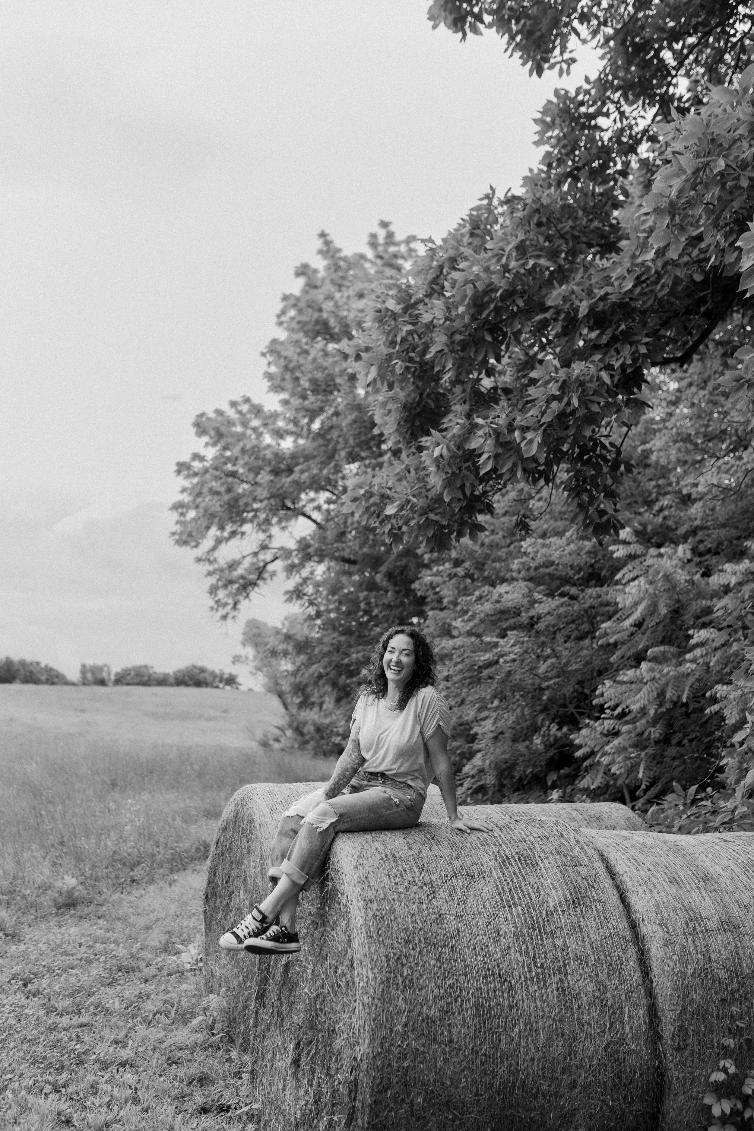 A woman with curly hair and tattoos, sitting on top of a hay bale in a field, smiling and looking to the side, with trees and an overcast sky in the background.