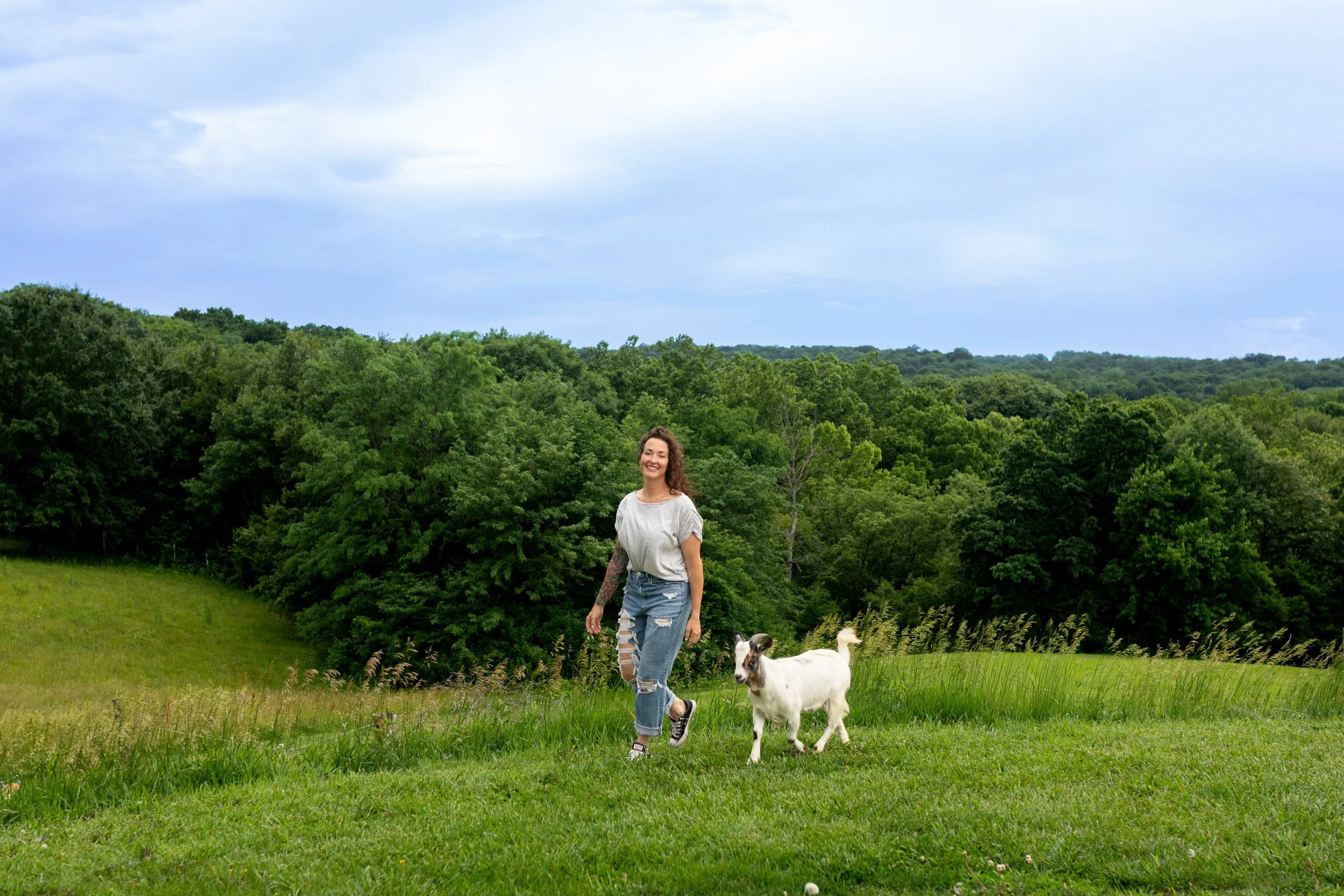 A woman walking with two goats on a grassy area with trees and a cloudy sky in the background.
