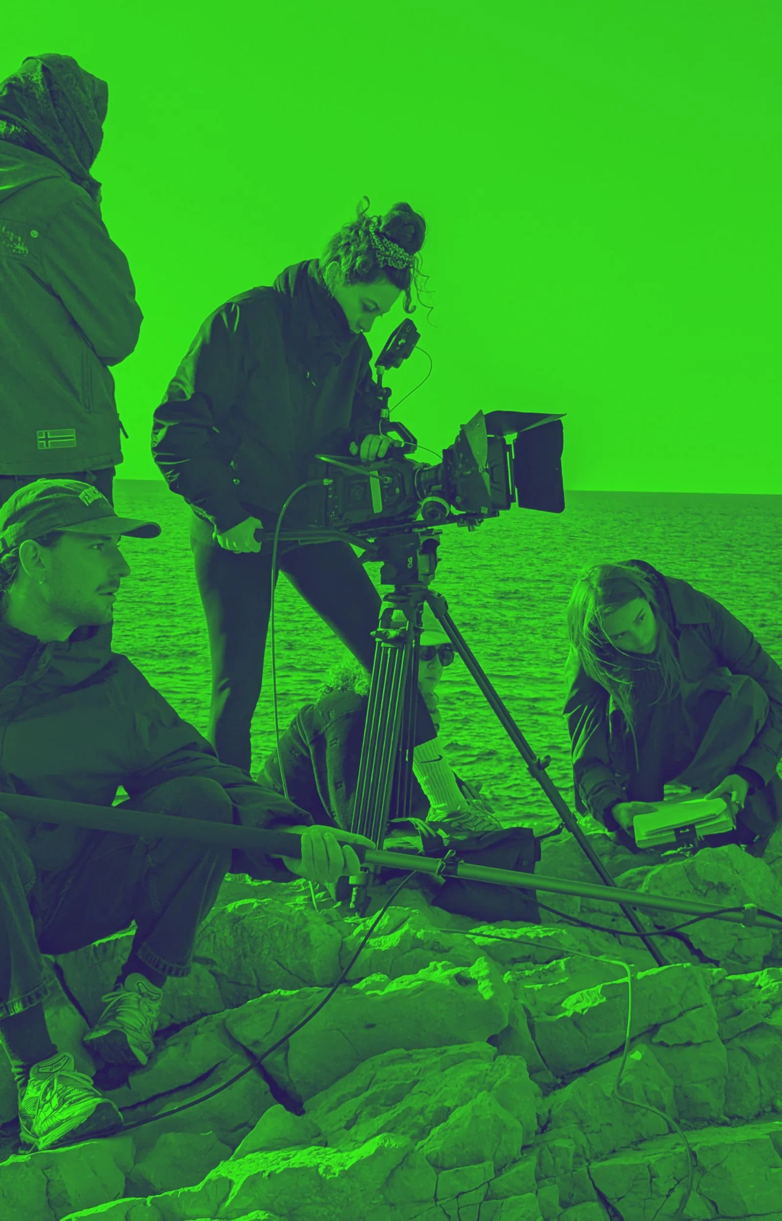 Une équipe de production filme une scène près de la mer par une journée venteuse, certains membres travaillent avec des équipements de tournage, d'autres prennent des notes ou observent, tous portent des vêtements chauds et des casquettes.
