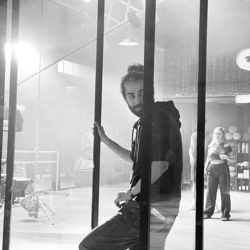 Un jeune homme avec une barbe et des cheveux bouclés derrière une paroi en verre, dans un studio ou un espace créatif, avec d'autres personnes en arrière-plan.