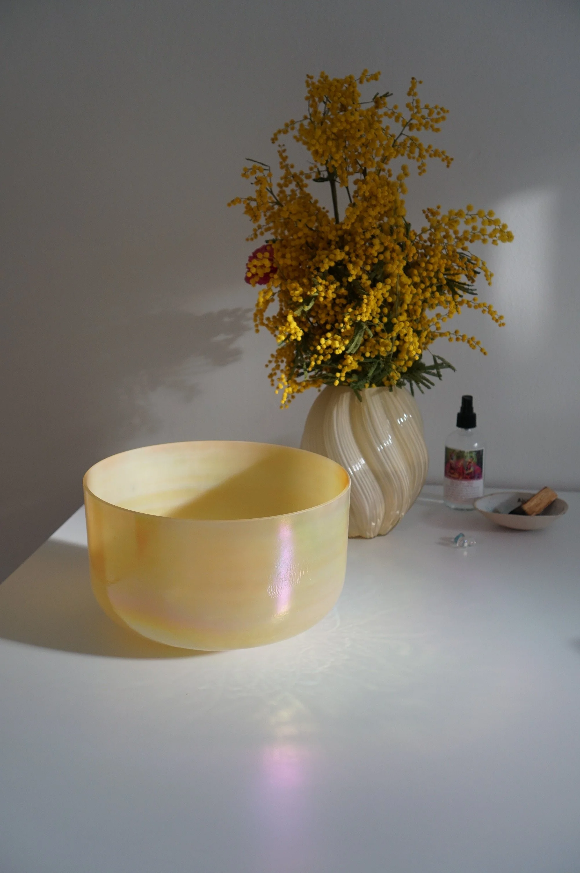 A crystal tones singing bowl with a yellow, iridescent bowl in the foreground, a beige vase with yellow and red flowers in the background, and a small spray bottle, dish with a wooden stir stick, and some jewelry on the right.