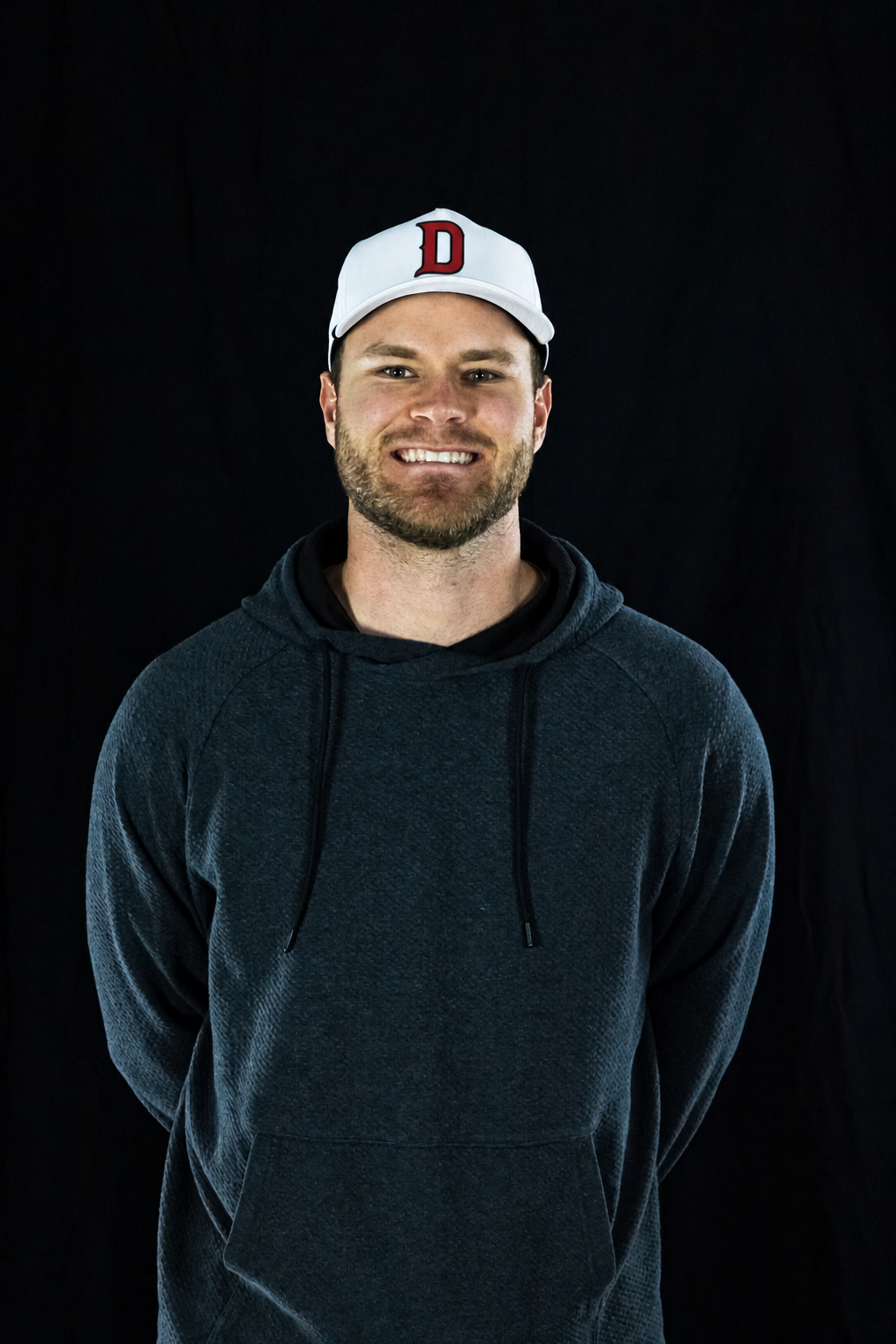 A smiling young man, wearing a white baseball cap with a red "D" on the front and a dark hoodie, standing against a black background.