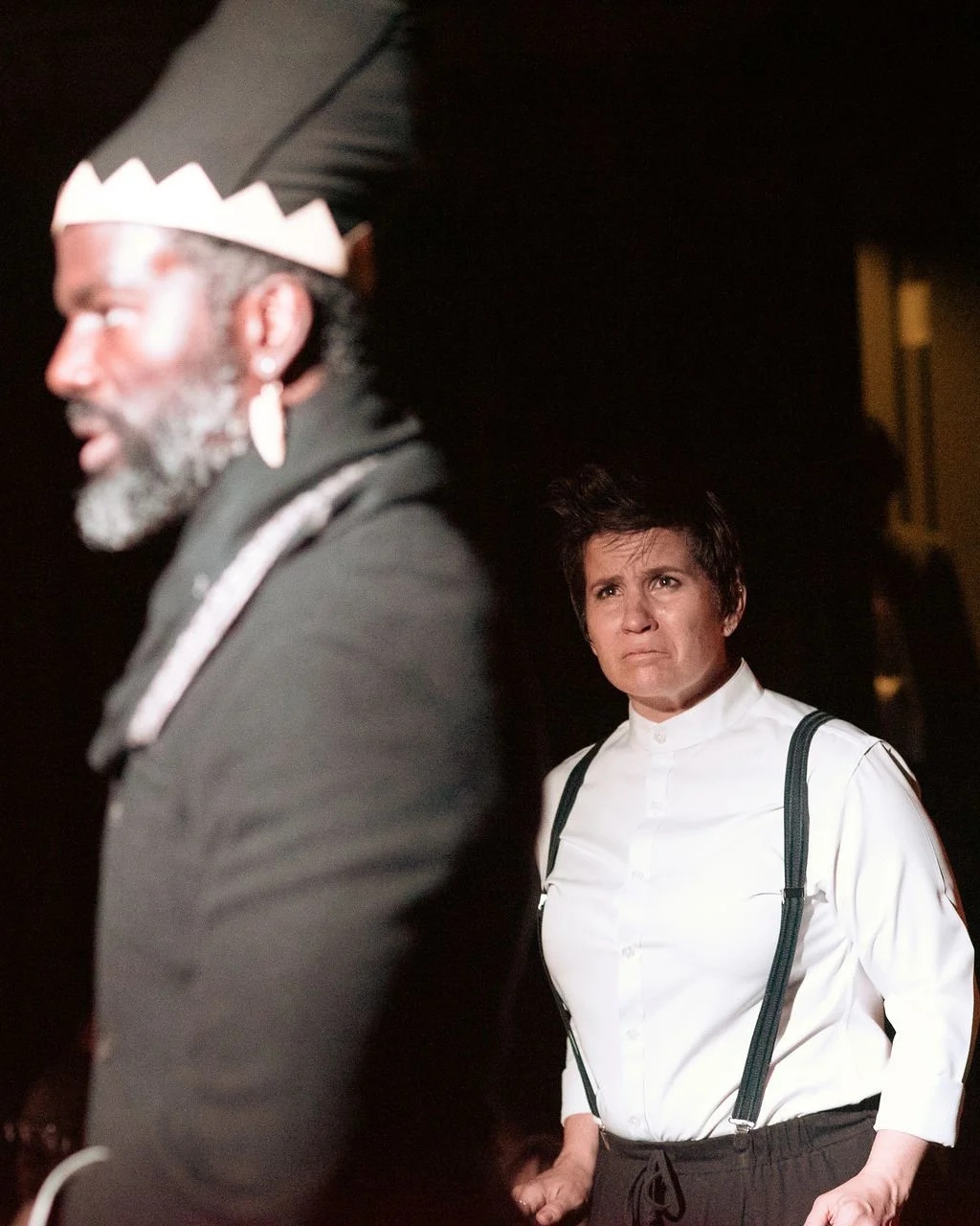 A woman with short dark hair looks upset or angry while standing indoors near a dark wall, wearing a white button-up shirt and suspenders. Behind her, a man with a beard, earrings, and a black headpiece is blurred.