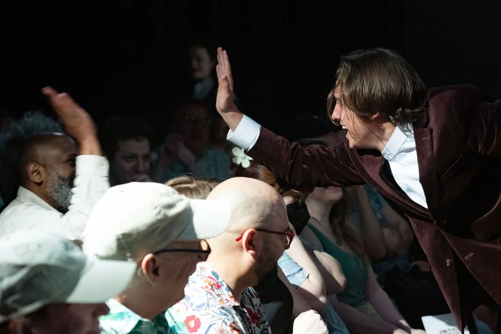 A man with shoulder-length hair high-fives a seated group of diverse people at an indoor event, with other attendees visible in the background.