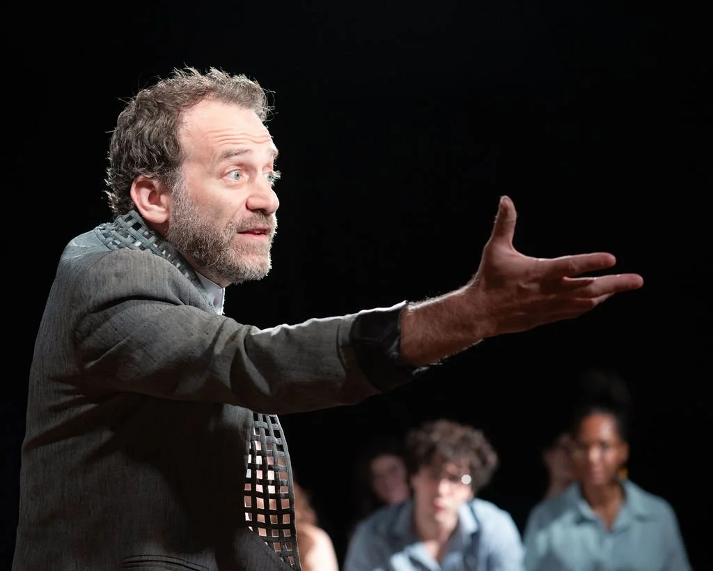 A man with curly hair and a beard gestures with his arm extended while speaking to an audience. Several people are seated behind him, listening.
