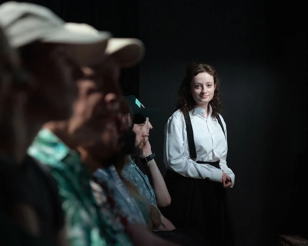A young woman standing in focus with others seated in front of her, in a dimly lit setting with a dark background.