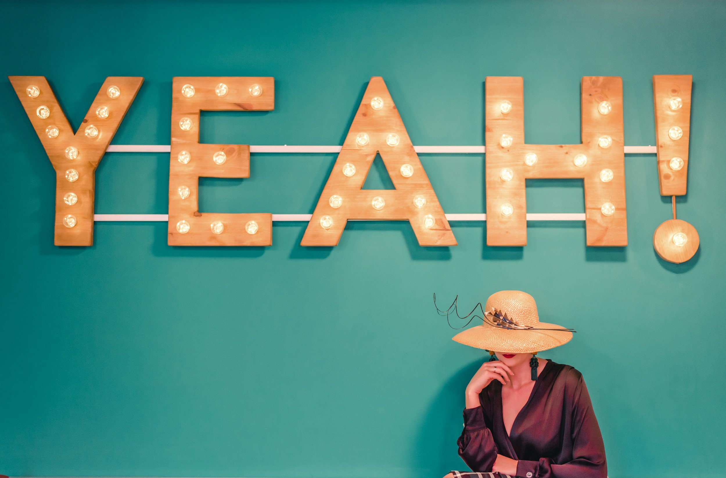Party decor featuring a large wooden sign that spells 'YEAH' with light bulbs, against a teal wall, and a woman wearing a wide-brim straw hat and a black sheer dress, with her hand near her face.