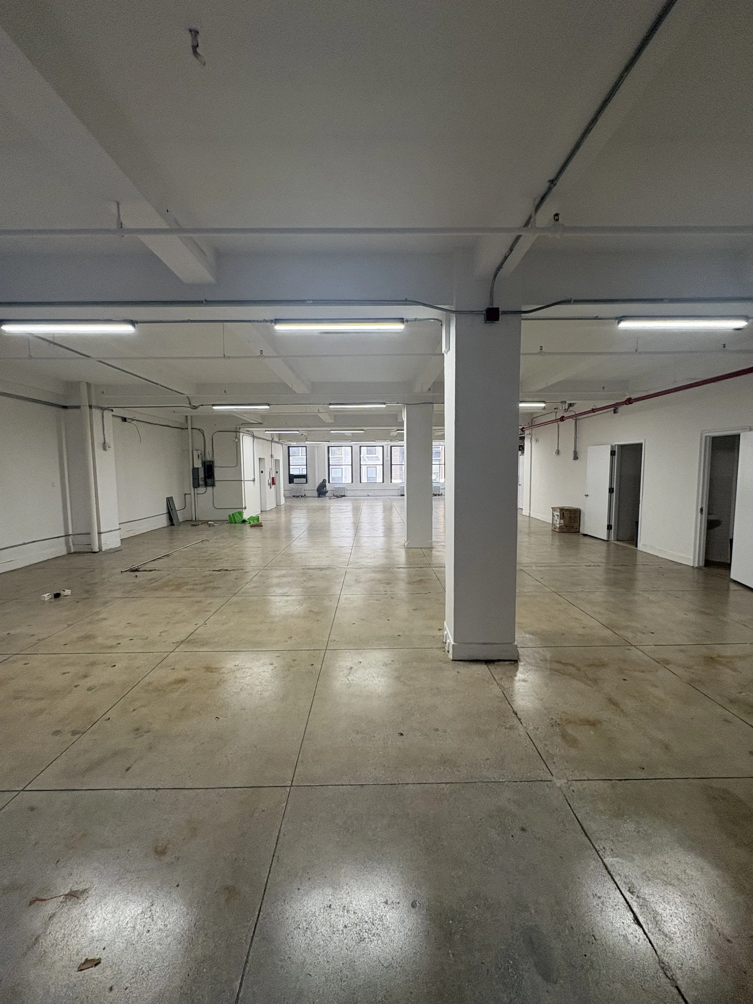 Empty interior space with concrete floor, white walls, support columns, fluorescent ceiling lights, and a large window at the far end.