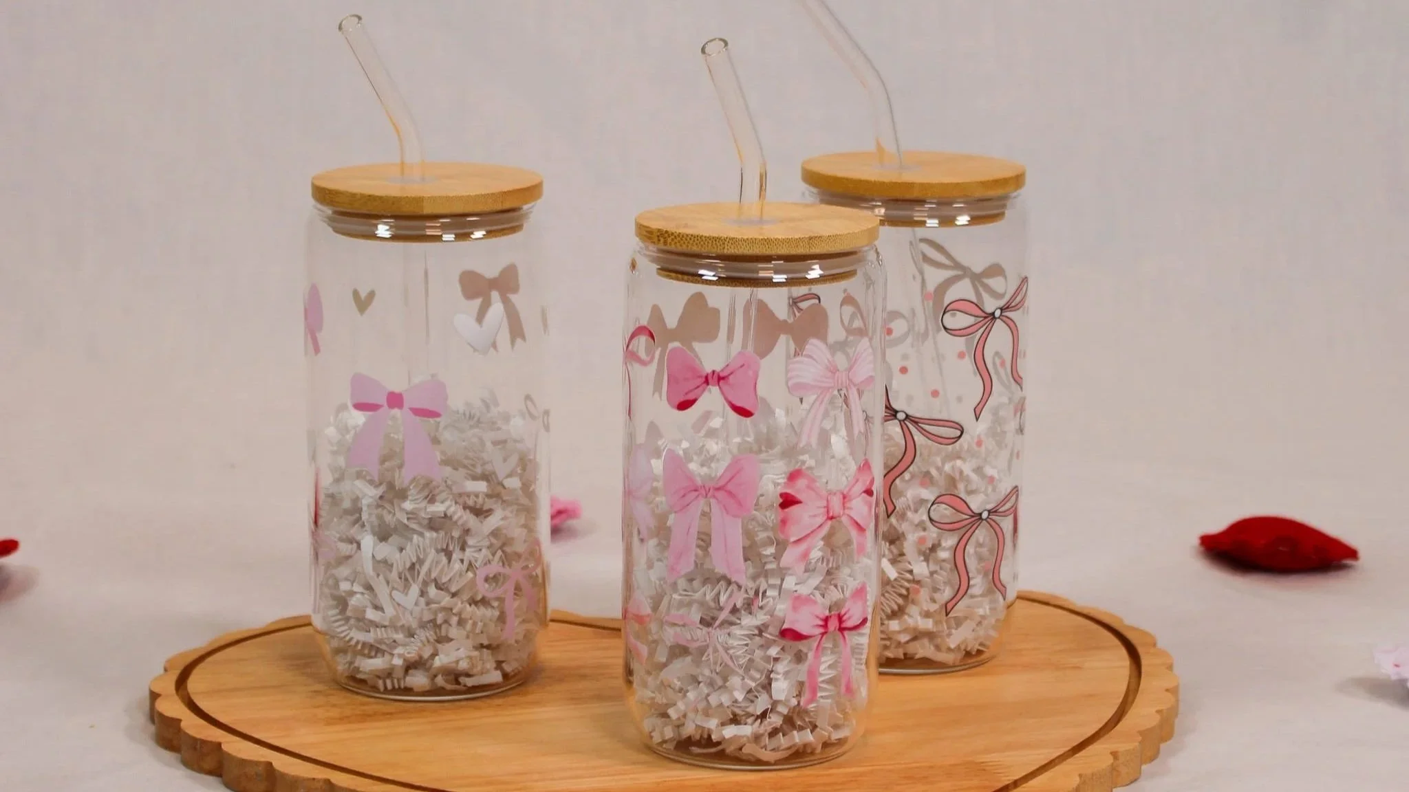 Three glass jars with wooden lids and pink ribbon and bow decorations, filled with shredded paper, on a round wooden tray.