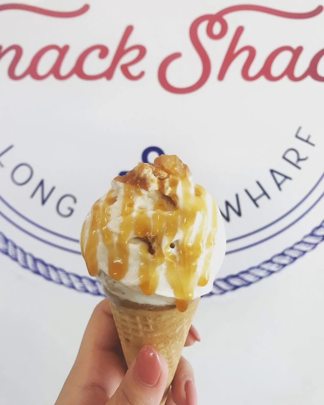 A vanilla ice cream cone topped with caramel sauce and chopped nuts held by a person's hand in front of a white wall with a red and dark blue logo that says 'Snacks' and 'Chow'.