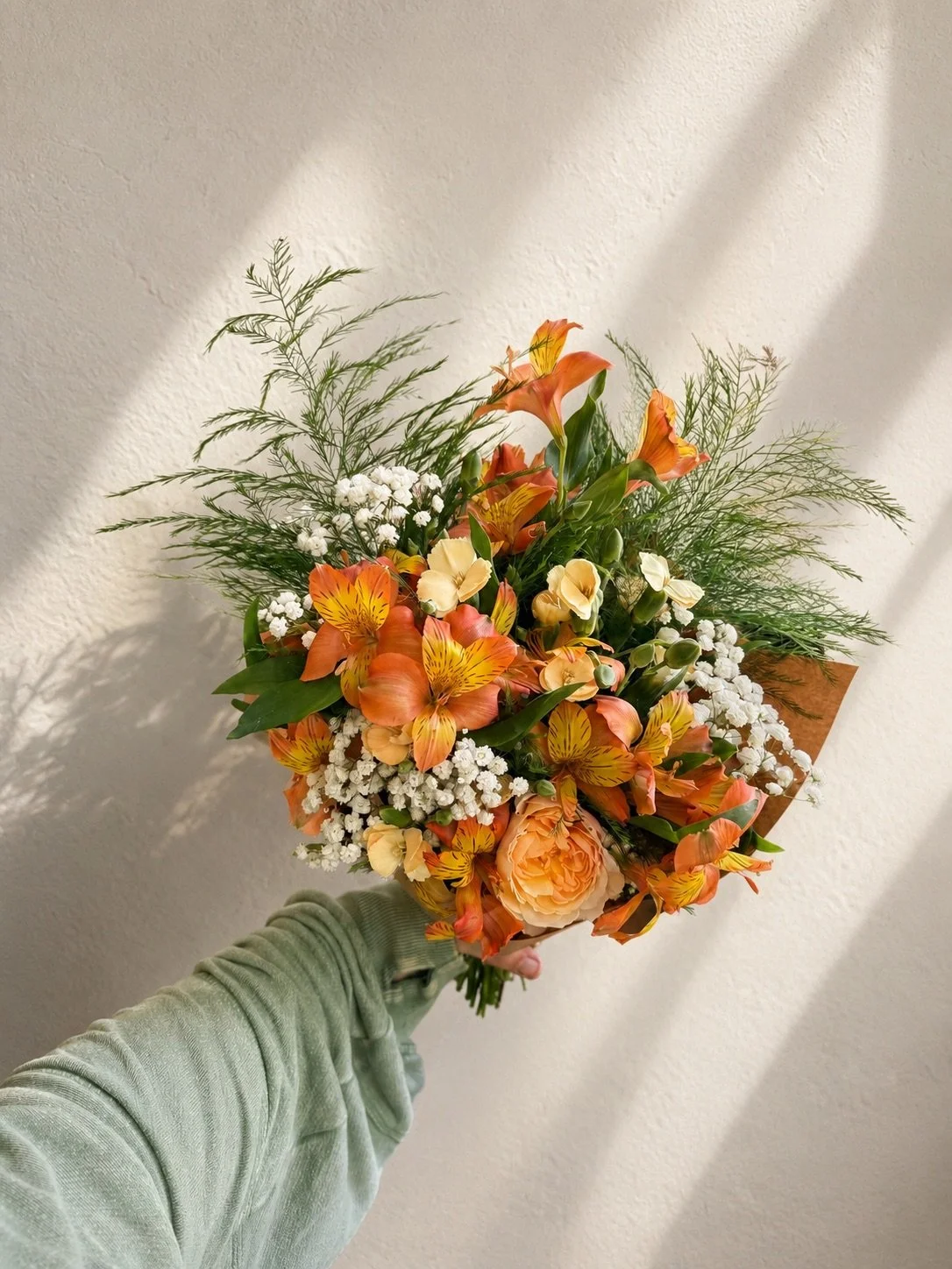 Hand holding a bouquet of orange lilies, peach roses, white baby's breath, and green foliage against a white wall.