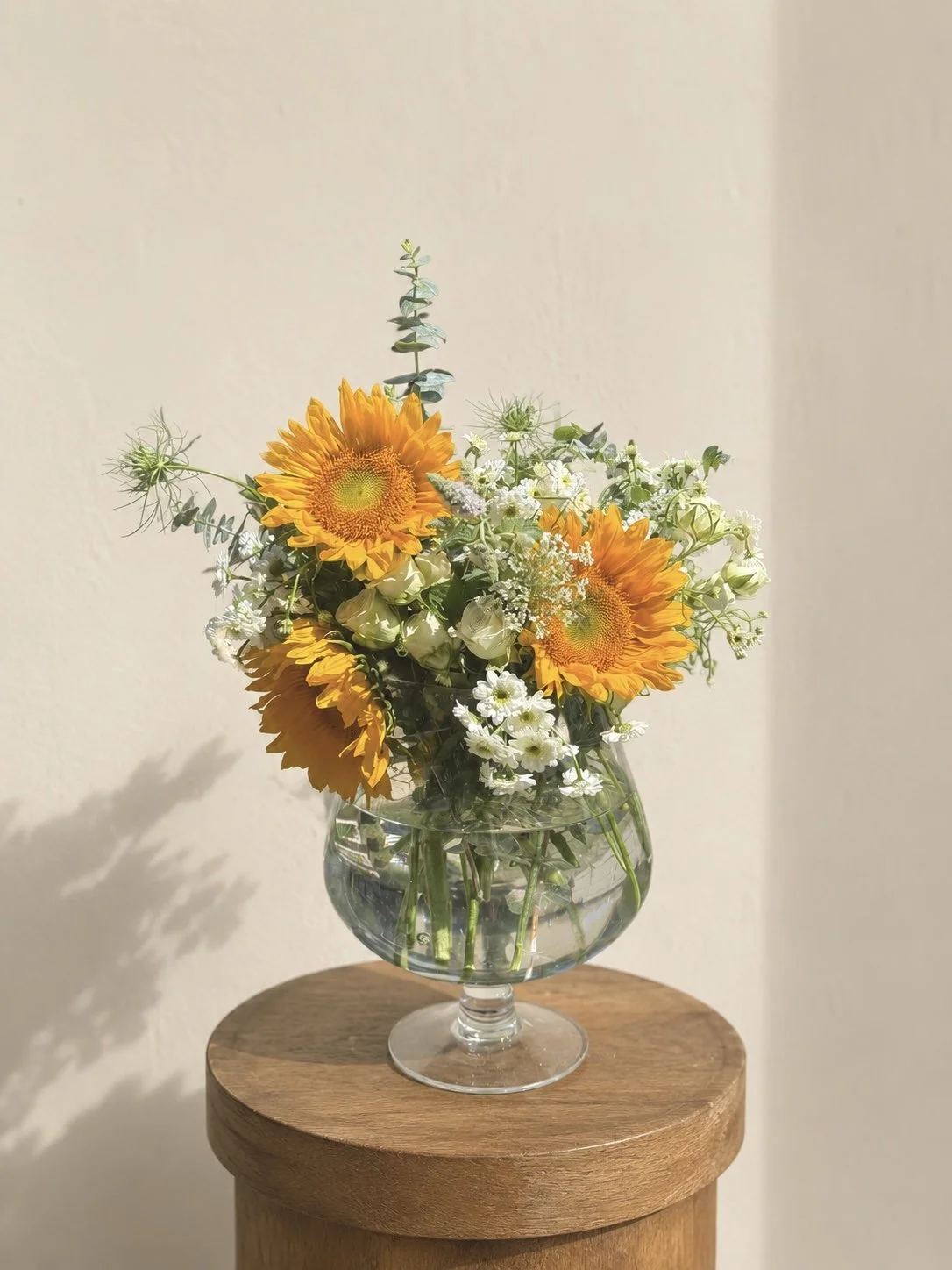 A glass vase with water holding a bouquet of sunflowers, white roses, small white flowers, and greenery, placed on a wooden stool against a plain wall.