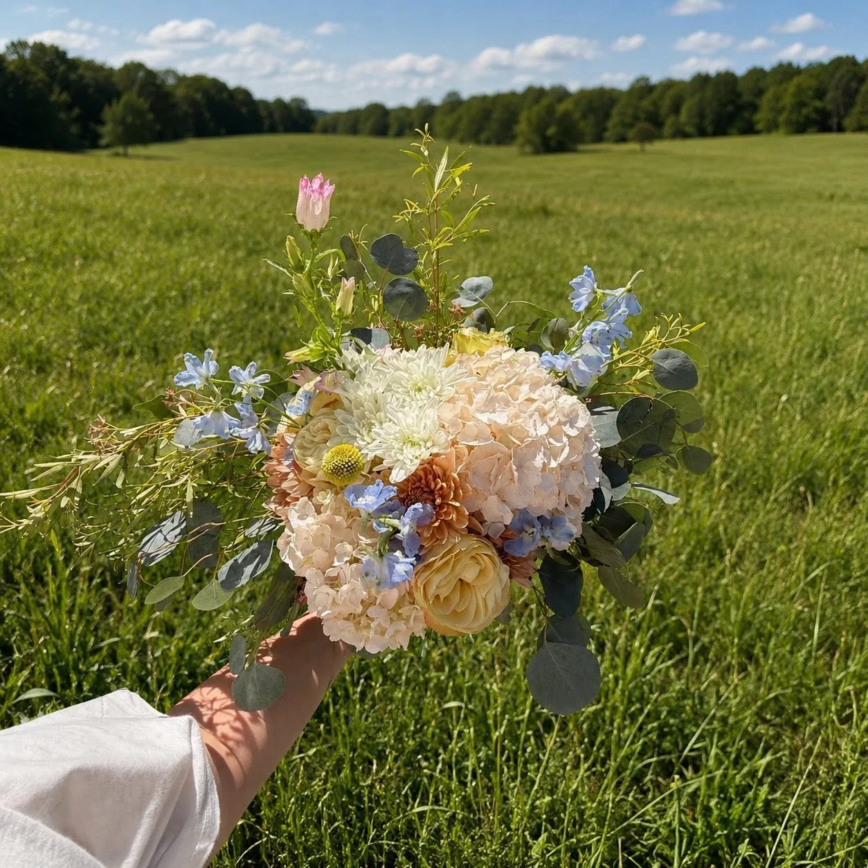 Person holding a bouquet of pastel-colored flowers in a green field on a sunny day with a blue sky and clouds.