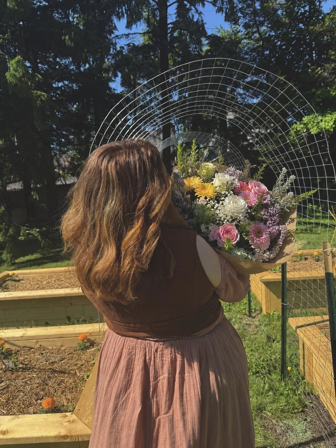A woman with long, wavy, light brown hair holding a large bouquet of mixed flowers outdoors in a garden with raised beds and trees.