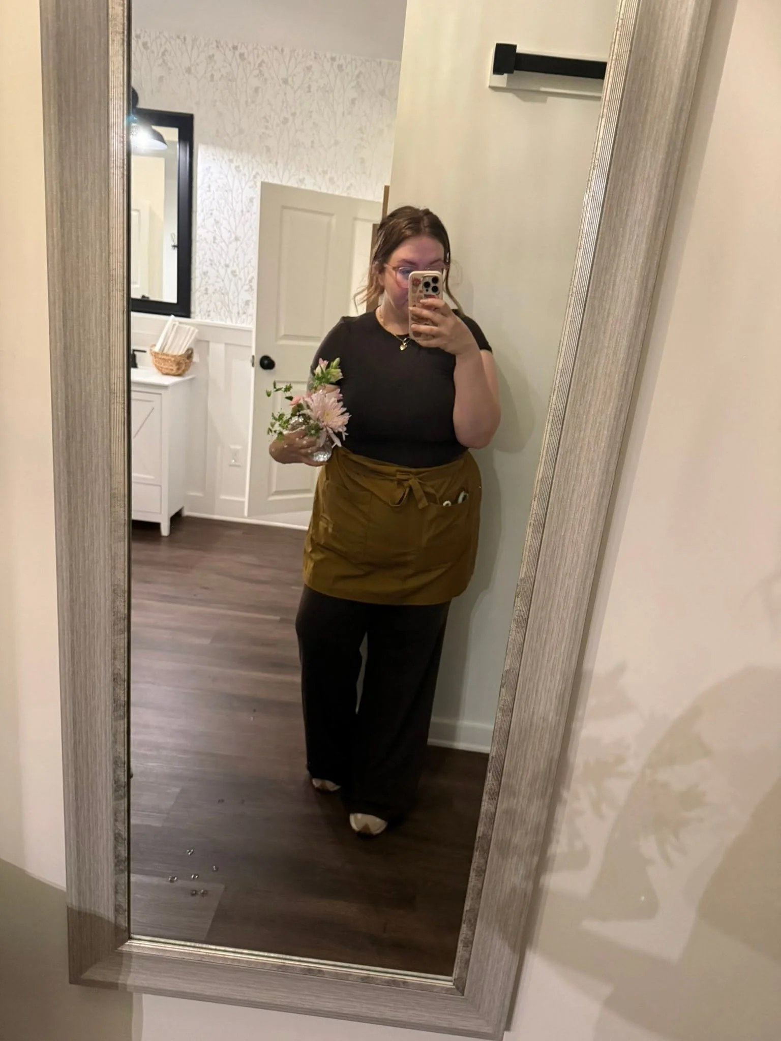 Woman taking a mirror selfie holding a bouquet of flowers, wearing glasses, black top, and brown skirt, in a room with wooden floors and white walls.