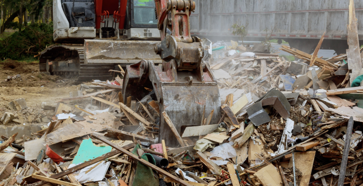 Heavy construction machinery clearing debris in a demolition site with scattered wood, paper, and other materials.