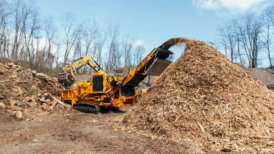 A yellow and black wood chipper processes a large pile of wood chips at a recycling site.