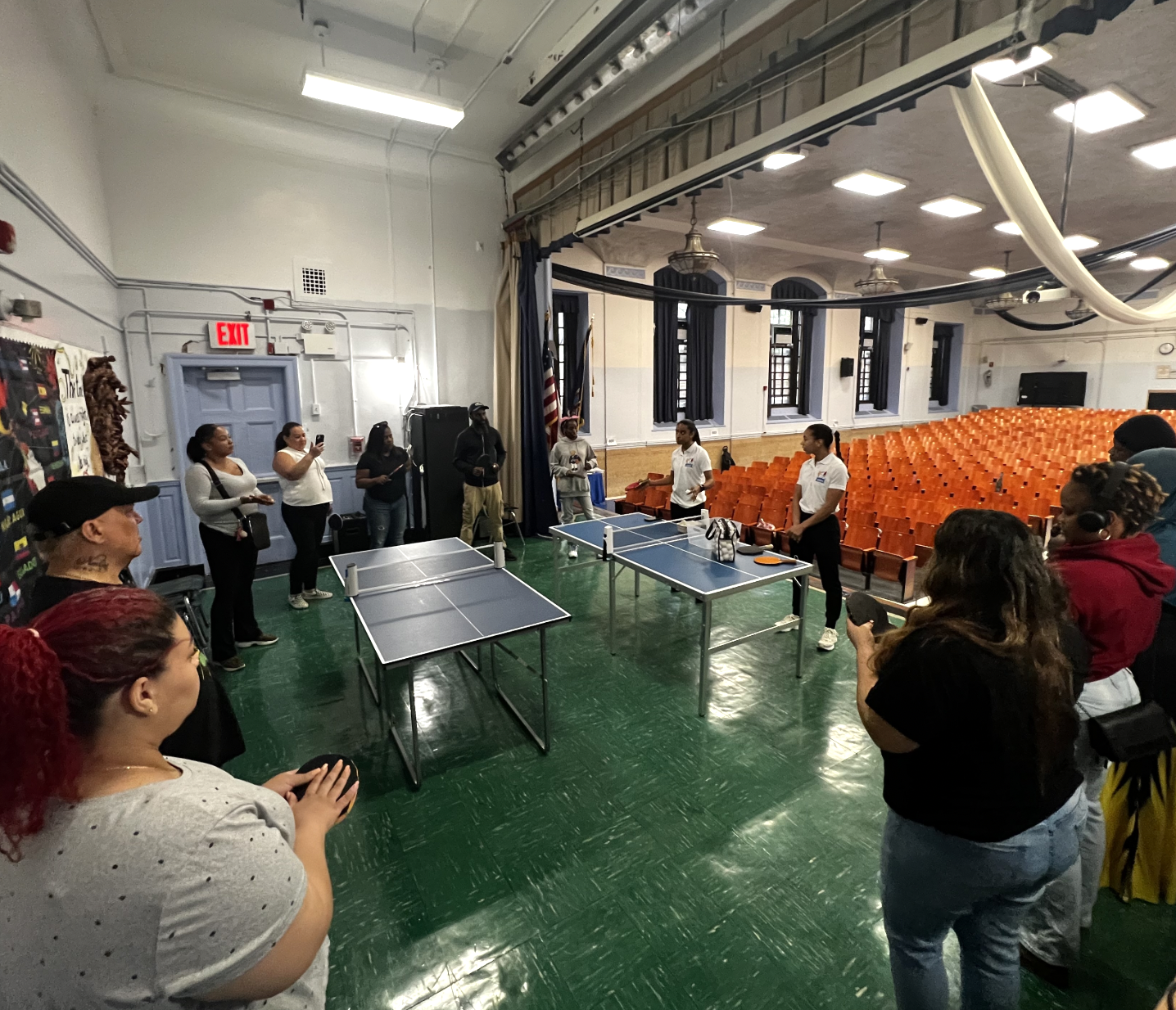 A group of people watching a ping pong game in a school auditorium with orange seats and high ceilings.
