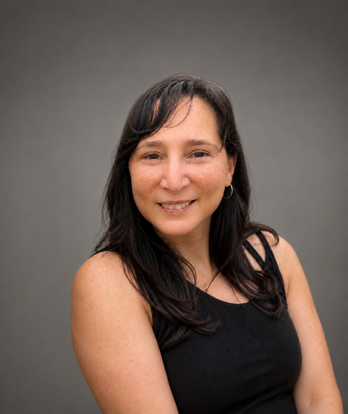 A woman with dark hair and freckles, smiling, wearing a black sleeveless top and gold hoop earrings, standing against a plain gray background.