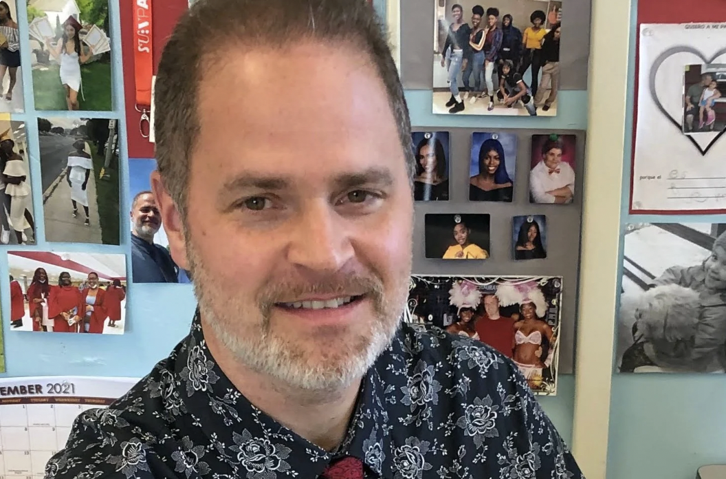A smiling man with a beard and short hair takes a selfie in front of a wall decorated with various photographs and posters of people in different settings.