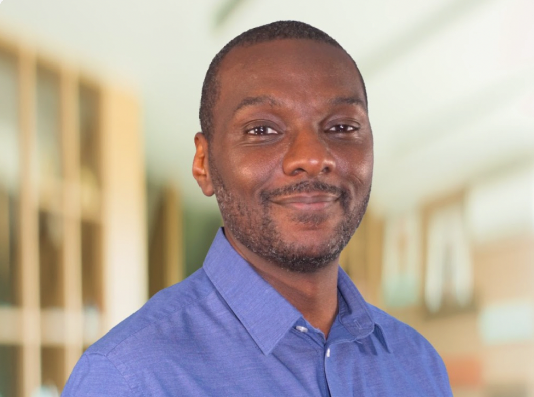 A smiling man with short hair and a beard, wearing a blue button-up shirt, standing indoors with blurred background of bookshelves and windows.