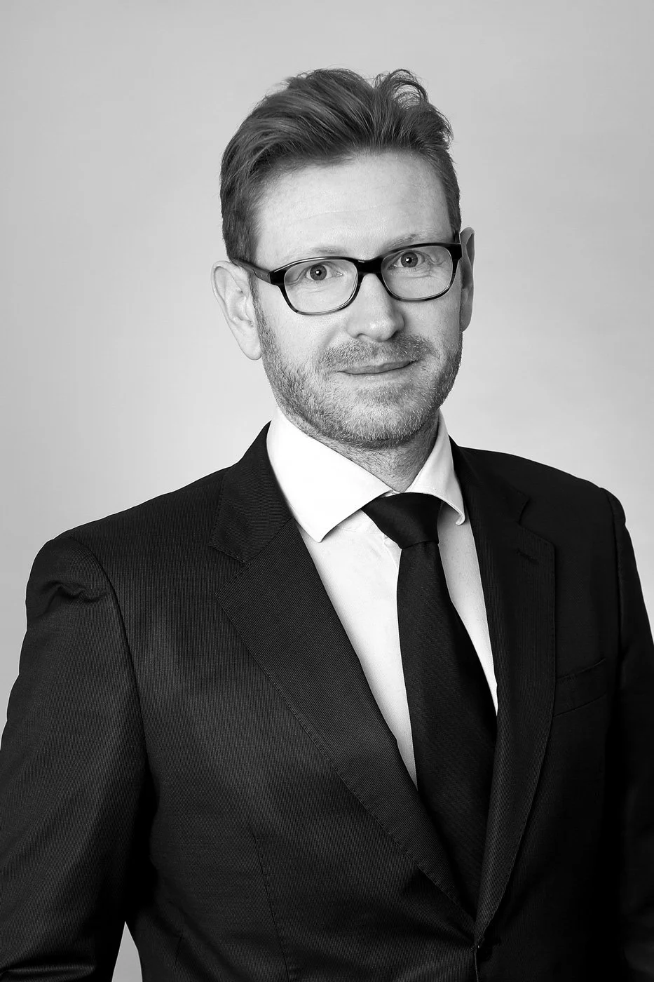 Black and white professional headshot of a man wearing glasses, a suit, white shirt, and tie, looking at the camera with a slight smile.