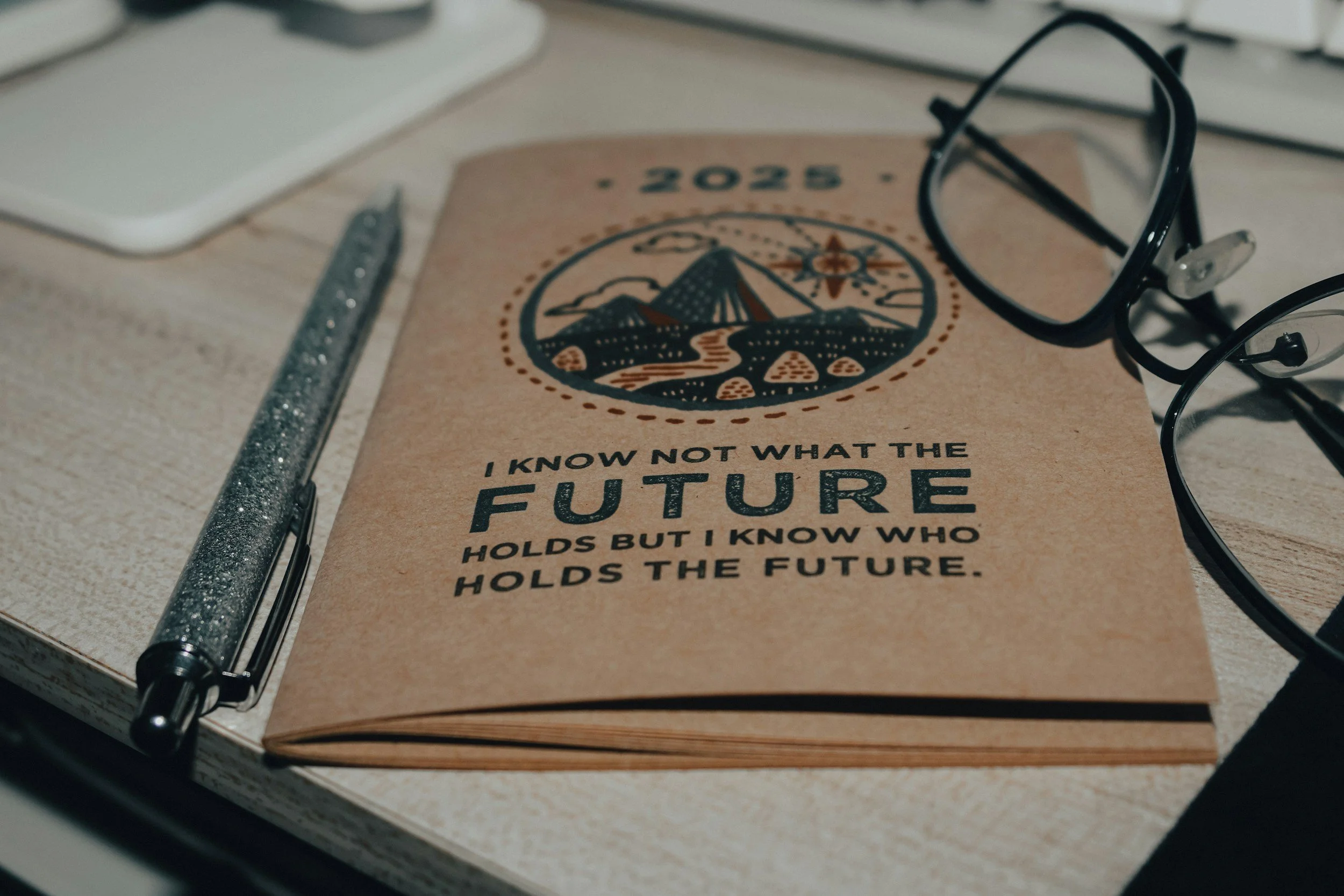 A booklet titled '2023' with an illustration of a mountain, river, and compass on the cover. The quote, 'I know not what the future holds but I know who holds the future,' is visible. A pen and a pair of glasses rest on the booklet, on a desk near a computer.