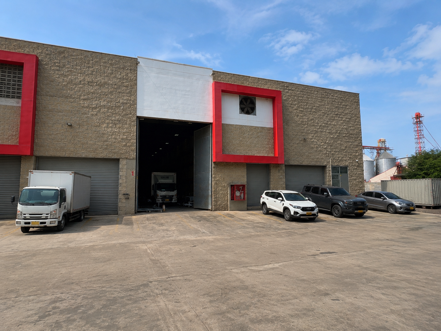 Fachada de un edificio industrial con varias entradas para camiones y autos estacionados delante, cielo despejado, en el fondo se ven estructuras metálicas y tanques de almacenamiento.