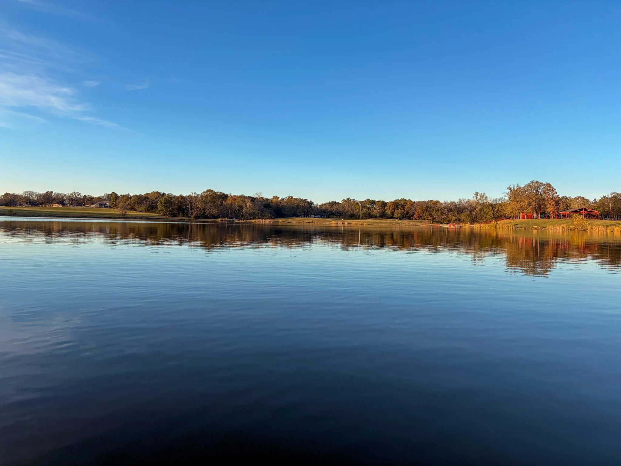 A lake reflecting the blue sky and trees along the shoreline, with a clear sky and some wispy clouds.
