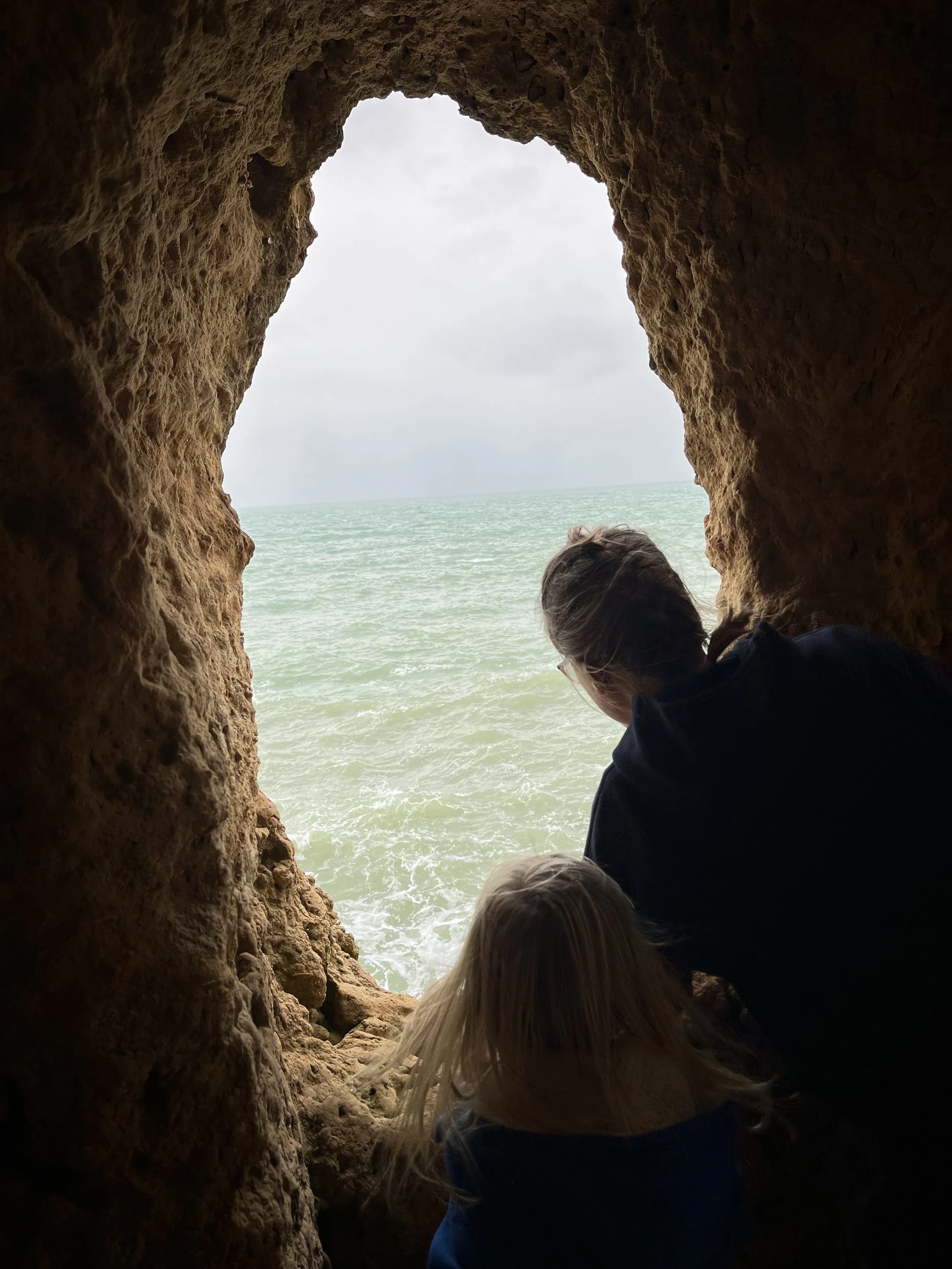 View from inside a small cave or rock formation looking out towards the ocean. The scene includes sandy-colored textured rocks framing the opening and a person with blonde hair looking out at the water.”}