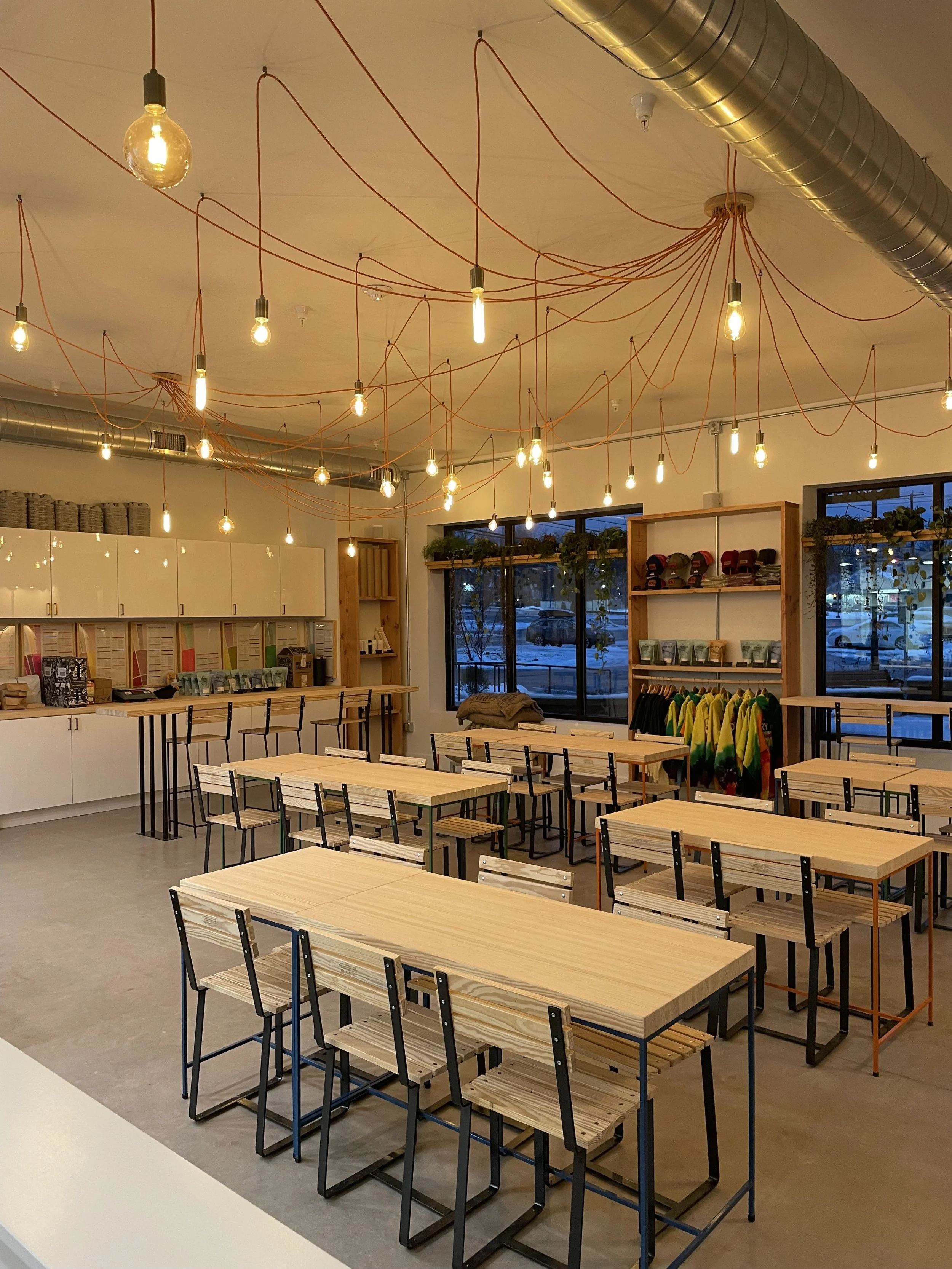 Interior of a modern, minimalist cafe or co-working space with wooden tables and black metal chairs. Ceiling overhead lighting includes exposed filament bulbs with orange cords and large silver ventilation duct work. Large windows reveal a winter scene outside.