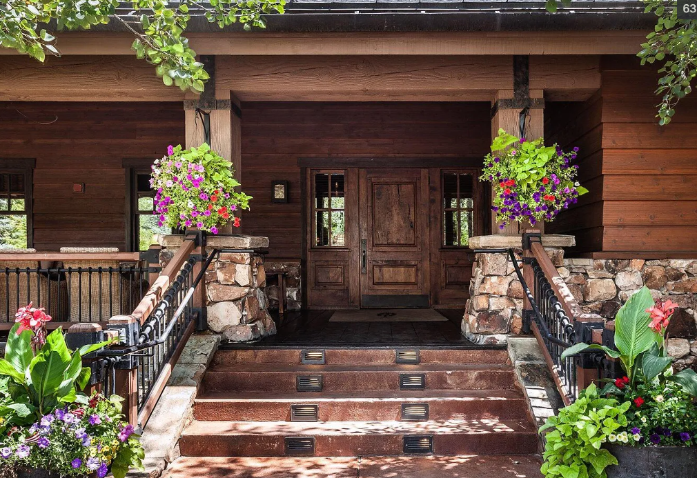 Front porch of a wooden house with stone accents, featuring hanging flower baskets and potted plants with colorful flowers.