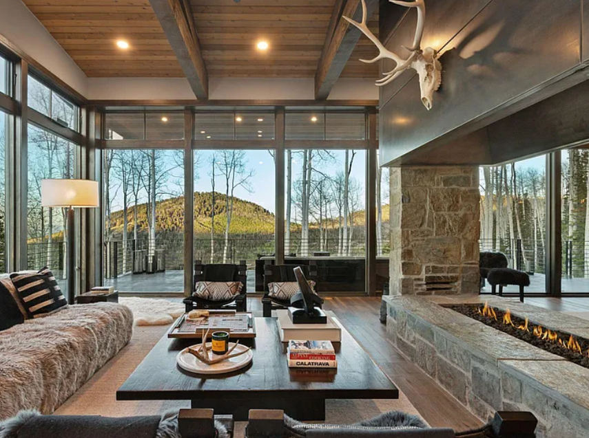 Living room with large glass windows overlooking a mountain and trees, stone fireplace, deer skull with antlers hanging on wall, modern furniture, and wooden ceiling.