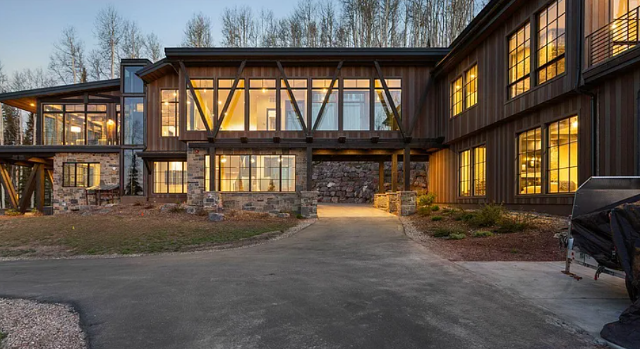Modern two-story house with large glass windows and a stone foundation, illuminated from inside during twilight, surrounded by trees and a paved driveway.