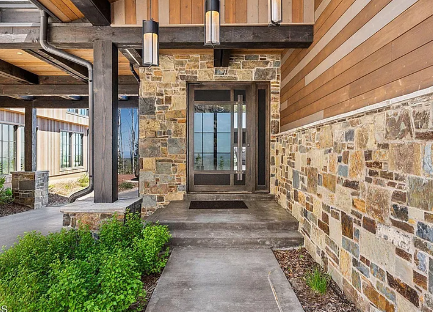 View of a building entrance with a glass door, stone and wood siding, and a concrete sidewalk.
