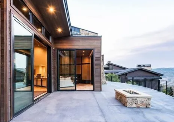 Modern house with large glass sliding doors and a spacious outdoor patio with a fire pit, overlooking a scenic landscape.