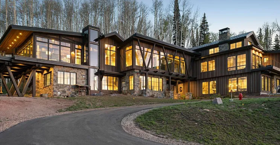 Modern multi-story house with extensive glass windows, wooden siding, and stone foundation, situated on a sloped driveway with trees in the background, illuminated from inside at dusk.