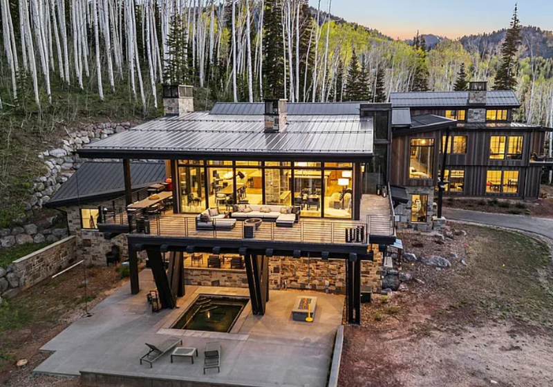 A modern house built on a hill surrounded by trees, with large glass windows, a wooden deck, and outdoor seating area, during dusk.