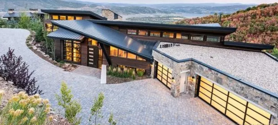 Modern house with angular design, stone and wood exterior, large garage doors, surrounded by landscaped yard and overlooking a scenic landscape.