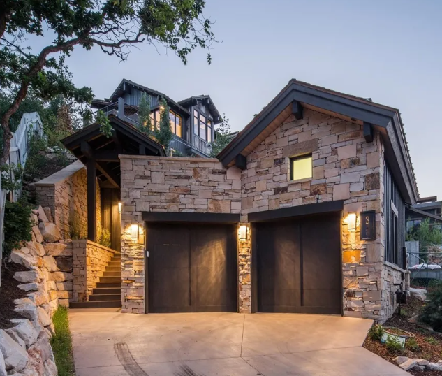 A modern house with stone exterior, two garage doors, and a stone staircase leading up to an entrance, surrounded by trees and landscaping at dusk.
