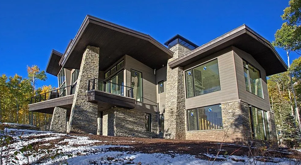 Modern house with large windows, stone and wood exterior, surrounded by trees, with a clear blue sky.